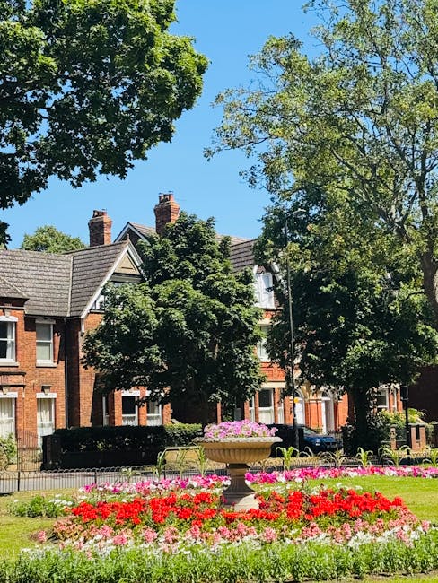 This image features a vibrant flower bed in a park setting with a central stone planter filled with pink flowers, surrounded by neatly arranged red, pink, and white blooms. Tall green trees with lush 