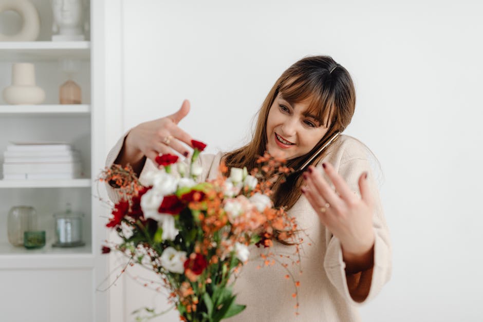 A woman with shoulder-length brown hair and bangs is smiling while holding a vibrant bouquet of fresh flowers, including red roses, white lisianthus, orange alstroemeria, and pink mimosa, wrapped in c