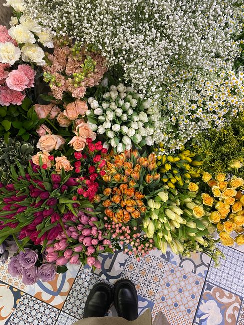 A vibrant and diverse floral display arranged on a patterned tiled floor, featuring a variety of fresh flowers including pink tulips, orange and yellow roses, white daffodils, purple roses, and small 