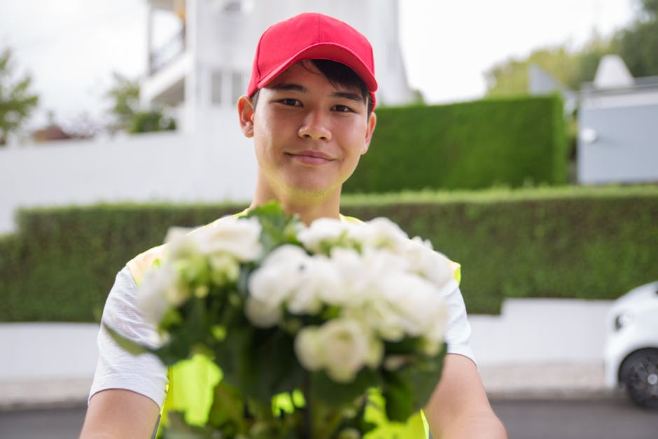 A male flower delivery courier wearing a red cap and a high-visibility vest is standing outdoors in a residential area, presenting a vibrant bouquet of white roses and green foliage. The bouquet is wr