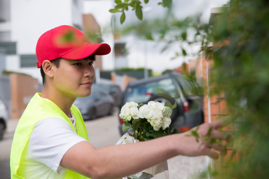 A young male flower delivery courier wearing a red cap and a high-visibility yellow vest is shown handing over a bouquet of fresh, white hydrangeas wrapped in white paper to a customer. The bouquet is
