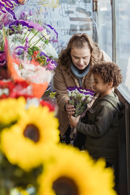 A woman and a young girl in a flower shop with large windows, exchanging a bouquet of purple and white flowers wrapped in purple paper, surrounded by vibrant floral arrangements including yellow sunfl