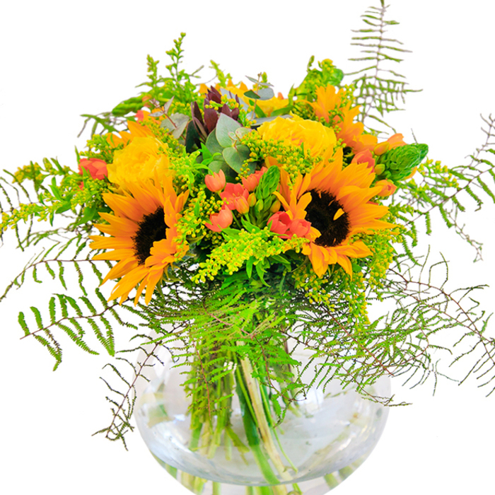 From the first glance, this bouquet radiates the joyful energy of Wood Green in peak season. Sunflowers and golden roses bloom together, their petals gracefully layered to capture every ounce of available light-a vision reminiscent of a sun-dappled spot on Ducketts Common. The composition abounds with life: sleek, leafy greens and delicate ferns arc outward, mingling with airy green sprigs and slender, upright spikes that add texture and fullness. Glossy clusters of scarlet hypericum berries sparkle unexpectedly against the soft, glowing blooms and matte textures, creating a vivid visual rhythm that dances through the bouquet. Hints of muted blue-green eucalyptus nestle quietly among brighter greens, balancing the arrangement with a rustic, natural air. Every stem is cradled by a round, clear glass vase, showcasing the patient craftsmanship from local florists and highlighting the tangle of freshly trimmed stems in the cool, clear water below. The gentle scent is more suggestion than scent-a blend of sun-touched rose and green, perfect for refreshing the air in any home. Ideal for milestone birthdays, heartfelt congratulations, or simply bringing cheer to a grey day, this bouquet brings the vivacity and warmth of Wood Green indoors, celebrating life's special moments in full bloom.